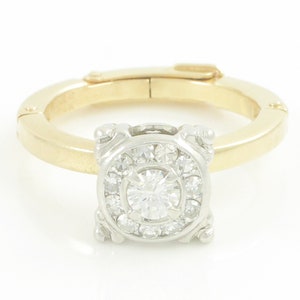 May include: A gold and white ring with a round diamond center stone surrounded by smaller diamonds. The ring has a unique design with a square setting and a hinged band.