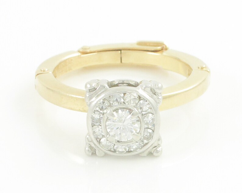 May include: A gold and white ring with a round diamond center stone surrounded by smaller diamonds. The ring has a unique design with a square setting and a hinged band.