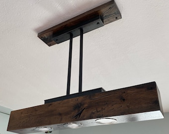 Op de afbeelding: Een rustieke houten lamp met een zwart metalen frame en drie ronde lampen. De lamp hangt aan het plafond en heeft een verweerde houten afwerking.