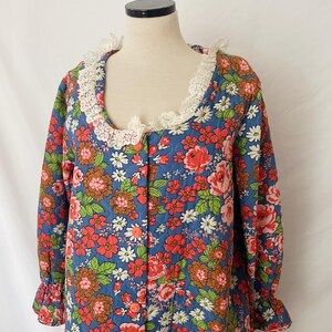 May include: A blue floral patterned robe with red, pink, white, and brown flowers. The robe has a white lace collar and ruffled sleeves. The robe is buttoned up and appears to be made of a quilted fabric.
