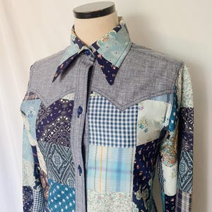 May include: A blue and white patchwork long-sleeve shirt with a pointed collar. The shirt is made of different fabrics with various patterns, including paisley, plaid, and gingham.