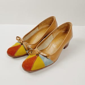 May include: A pair of vintage women's shoes with a tan leather upper and a colorful suede toe. The toe features a patchwork design in orange, yellow, and blue. Each shoe has a small bow detail and a low heel.