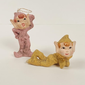 May include: Two vintage elf figurines. One is standing in a pink glittery outfit with a matching hat. The other is lying down in a gold glittery outfit with a matching hat. Both have painted faces with red lips and brown hair.