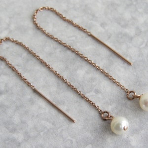 Può includere: Orecchini a filo in oro rosa con un delicato design a catena. Ogni orecchino presenta una barra dritta e una piccola perla bianca rotonda all'estremità, creando un look semplice ma elegante.