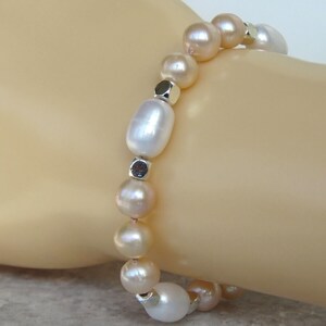 May include: A bracelet made of white and peach-toned pearls, with small silver-coloured metal beads. The pearls are different sizes and shapes, creating a unique design. The bracelet is displayed against a light background. The bracelet is approximately 18 cm in length.