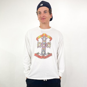 May include: White long-sleeve shirt with a Guns N' Roses graphic on the front. The graphic features a cross design with skulls and the band's name. The shirt is paired with black shorts and a navy baseball cap.