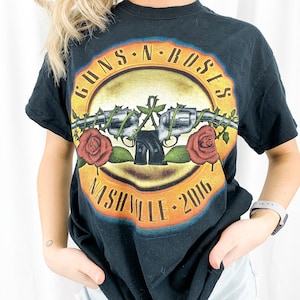 May include: Black t-shirt featuring a Guns N' Roses graphic. The design includes the band's name in gold lettering, crossed revolvers, red roses, and the text "Nashville 2016".