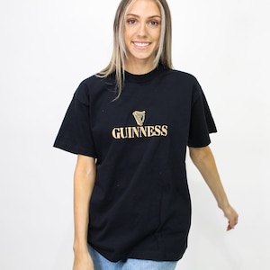 May include: Black t-shirt with the Guinness logo in gold. The logo features a harp symbol above the Guinness text. The shirt is a classic crew neck style.