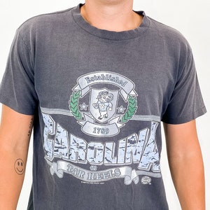 以下が含まれることがあります： グラフィックデザインが施されたチャコールグレーのTシャツ。「CAROLINA」の文字が青と白で描かれ、その上に「Established 1789」と書かれています。また、漫画のキャラクターと「TAR HEELS」の文字が入った紋章も付いています。