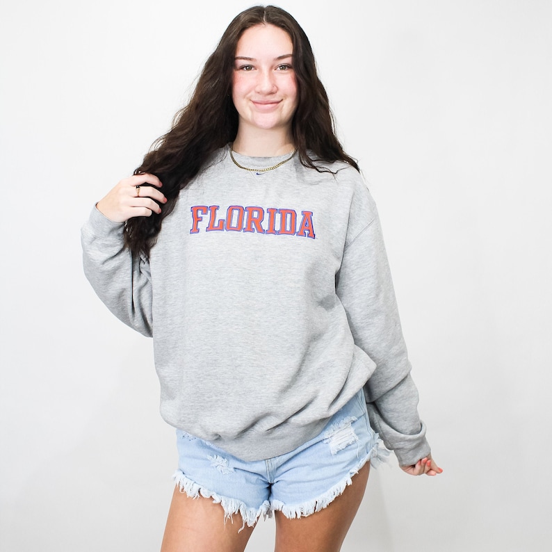 以下が含まれることがあります： 赤と青の文字で「FLORIDA」と書かれたグレーのスウェットシャツ。長袖のスウェットシャツはクルーネックです。その人は明るい青色のデニムショーツを履いています。