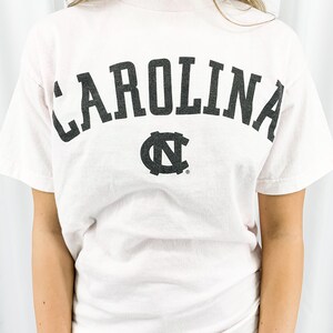 May include: A pale pink t-shirt featuring the word "CAROLINA" in large, black, arched lettering. Beneath the text is a black logo. The top has a crew neck and short sleeves.