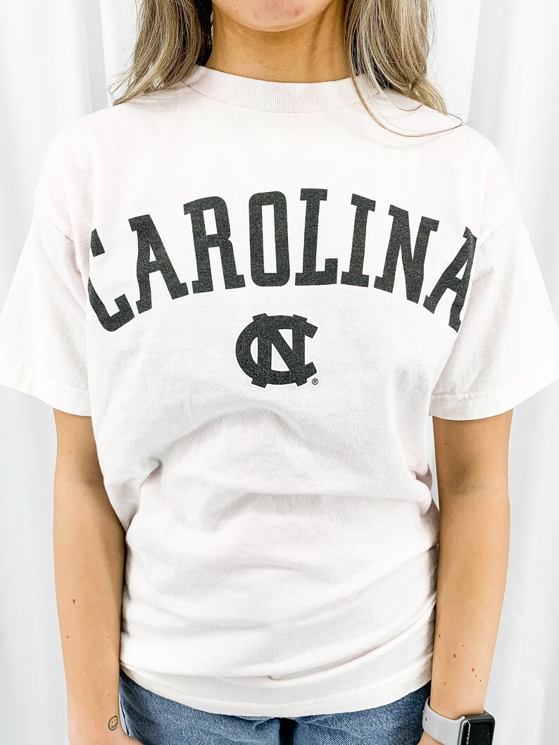 May include: A pale pink t-shirt featuring the word "CAROLINA" in large, black, arched lettering. Beneath the text is a black logo. The top has a crew neck and short sleeves.
