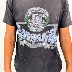 以下が含まれることがあります： 色あせたグラフィックが特徴の濃いグレーのTシャツ。「CAROLINA TAR HEELS」の文字と、雄羊のマスコットが付いたシールドデザインが描かれています。シャツには「Established 1789」の文字もヴィンテージスタイルで表示されています。