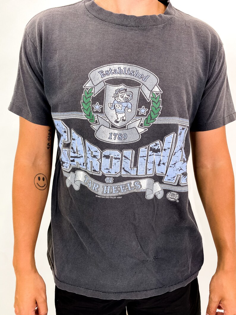 以下が含まれることがあります： 色あせたグラフィックが特徴の濃いグレーのTシャツ。「CAROLINA TAR HEELS」の文字と、雄羊のマスコットが付いたシールドデザインが描かれています。シャツには「Established 1789」の文字もヴィンテージスタイルで表示されています。