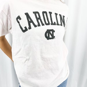 May include: A pale pink t-shirt featuring the word "CAROLINA" in large, dark grey letters. Beneath the word is a dark grey logo. The shirt is worn with blue jeans.