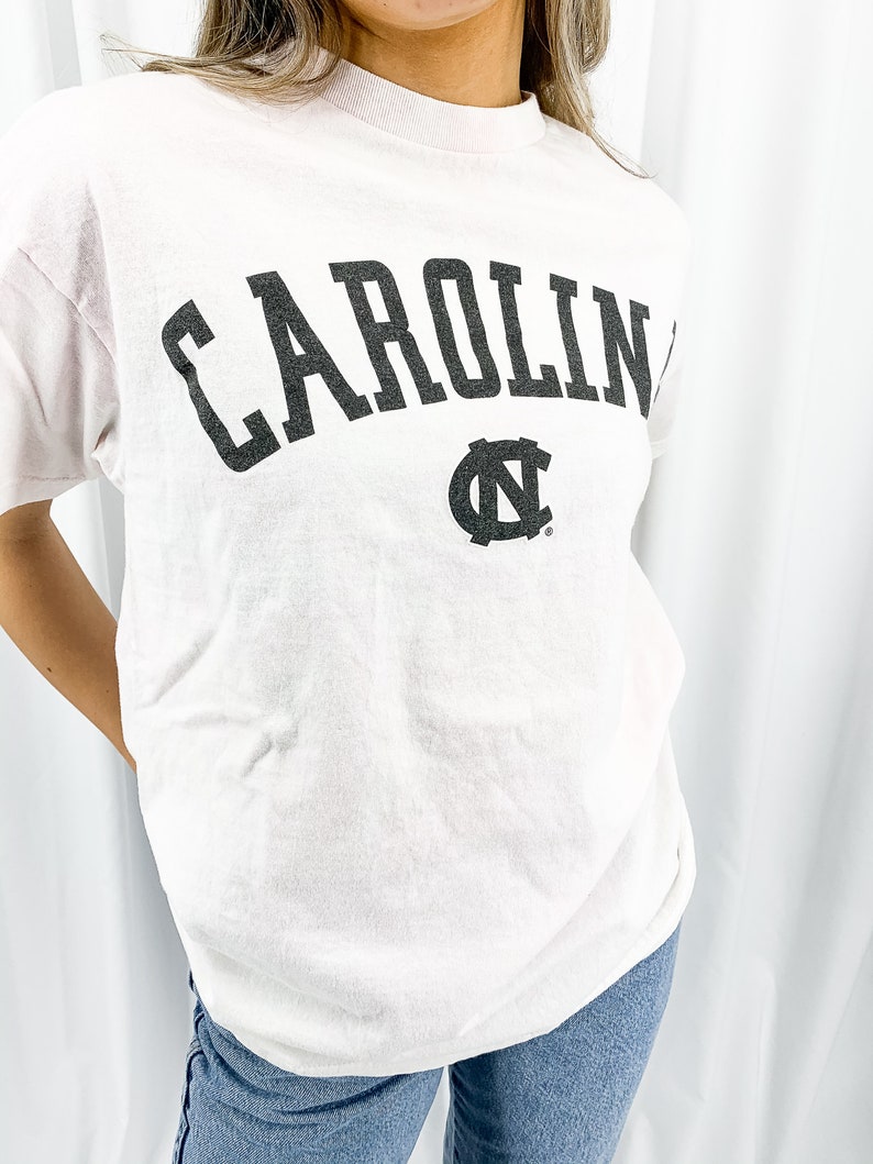 May include: A pale pink t-shirt featuring the word "CAROLINA" in large, dark grey letters. Beneath the word is a dark grey logo. The shirt is worn with blue jeans.