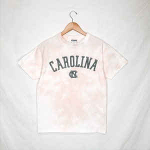 May include: A white t-shirt with a tie-dye effect in pink and peach hues. The word "CAROLINA" is printed across the chest in a distressed, dark grey font, with a matching logo below. The shirt is hanging on a wooden hanger.