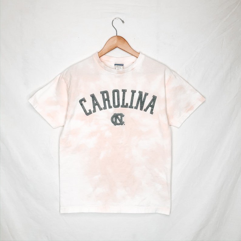 May include: A white t-shirt with a tie-dye effect in pink and peach hues. The word "CAROLINA" is printed across the chest in a distressed, dark grey font, with a matching logo below. The shirt is hanging on a wooden hanger.
