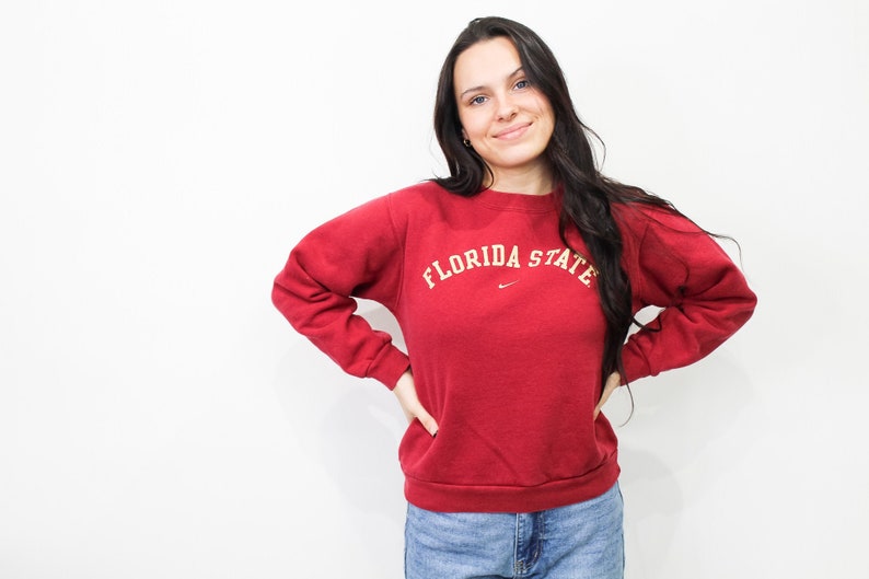 May include: A maroon crew neck sweatshirt with the text "FLORIDA STATE" in white arched lettering. A small Nike logo is visible below the text. The sweatshirt is worn with blue jeans.