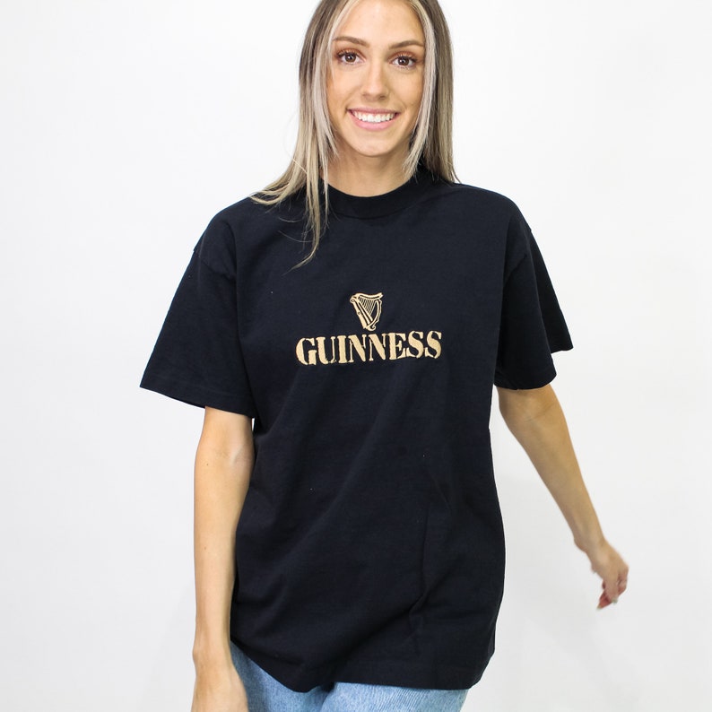 May include: Black t-shirt with the Guinness logo in gold. The logo features a harp symbol above the Guinness text. The shirt is a classic crew neck style.