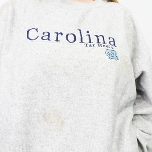 May include: A light gray sweatshirt with the word "Carolina" in navy blue script across the chest. Below the word "Carolina" is the text "Tar Heels" and a light blue logo. The sweatshirt has long sleeves.