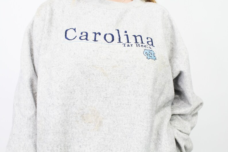 May include: A light gray sweatshirt with the word "Carolina" in navy blue script across the chest. Below the word "Carolina" is the text "Tar Heels" and a light blue logo. The sweatshirt has long sleeves.