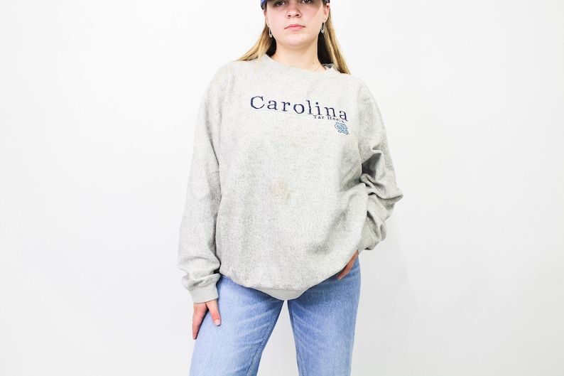 May include: A light gray sweatshirt with the word "Carolina" in navy blue script across the chest. Below the word "Carolina" is the text "Tar Heels" and a light blue logo. The sweatshirt is paired with blue jeans.