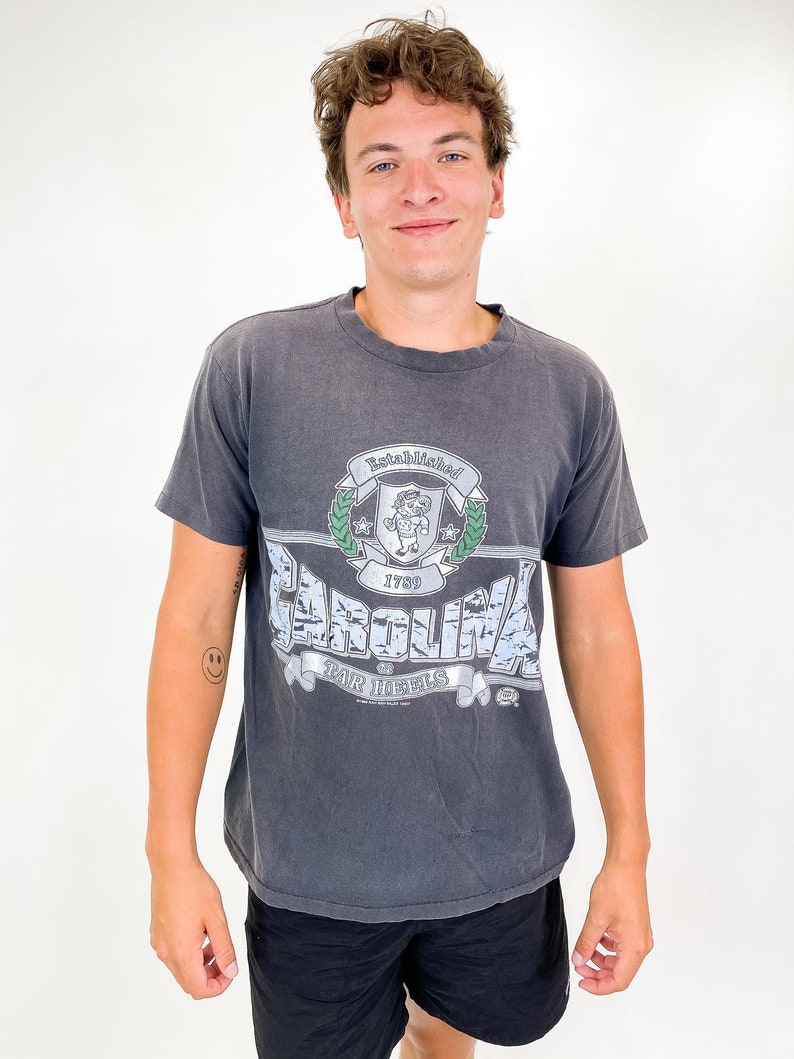以下が含まれることがあります： グラフィックデザインが施されたチャコールグレーのTシャツ。「CAROLINA」の文字が青と白で描かれ、その上に「Established 1789」と書かれています。また、漫画のキャラクターと「TAR HEELS」の文字が入った紋章も付いています。