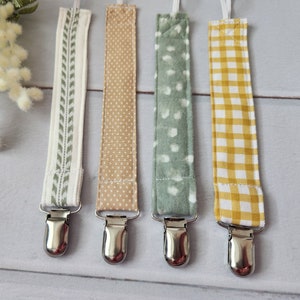 May include: Four baby pacifier clips with different fabric patterns. The patterns include a white and green leaf design, tan polka dots, white polka dots on green, and yellow gingham. Each clip has a silver metal clasp.
