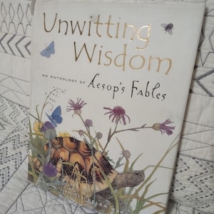 May include: A hardcover book titled "Unwitting Wisdom" with gold lettering and a colorful illustration of a tortoise, flowers, and butterflies. The book is an anthology of Aesop's Fables.
