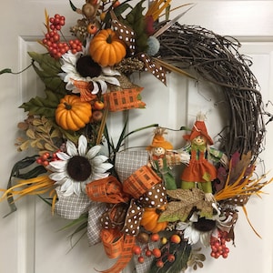 Op de afbeelding: Een herfstkrans met een basis van wijnstokken, versierd met oranje pompoenen, zonnebloemen, bessen en decoratieve vogelverschrikkers. De krans heeft bruine en witte stippenlinten en oranje geruite linten.