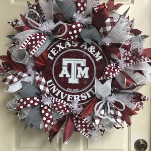 May include: A maroon and white wreath with a Texas A&M University logo in the center. The wreath is decorated with white polka dot ribbon, white and gray mesh, and white and red gingham ribbon.