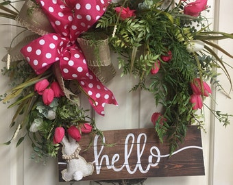 May include: A spring wreath with pink tulips, green leaves, and a pink and white polka dot bow. Two white fabric bunnies are attached to the wreath. A wooden sign with the word "hello" is attached to the bottom of the wreath.