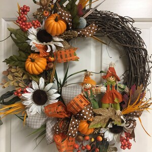Op de afbeelding: Een herfstkrans met een basis van wijnstokken, versierd met oranje pompoenen, zonnebloemen en bessen. Het heeft een grote strik met patronen en twee vogelverschrikkers. De krans is versierd met herfstbladeren.