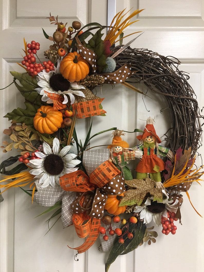 Op de afbeelding: Een herfstkrans met een basis van wijnstokken, versierd met oranje pompoenen, zonnebloemen en bessen. Het heeft een grote strik met patronen en twee vogelverschrikkers. De krans is versierd met herfstbladeren.