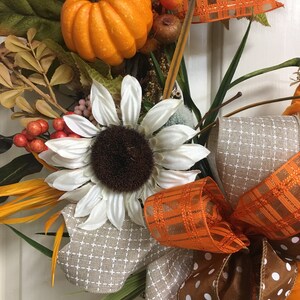 Op de afbeelding: Een herfstkrans met een feloranje pompoen, witte zonnebloem en diverse herfstbladeren. De krans bevat bruine en oranje decoratieve linten, met een ruit- en stippenpatroon. Bessen en andere kleine decoratieve elementen maken het ontwerp compleet.