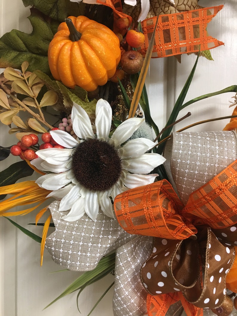 Op de afbeelding: Een herfstkrans met een feloranje pompoen, witte zonnebloem en diverse herfstbladeren. De krans bevat bruine en oranje decoratieve linten, met een ruit- en stippenpatroon. Bessen en andere kleine decoratieve elementen maken het ontwerp compleet.