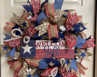 May include: A patriotic wreath with red, white, and blue ribbon, burlap, and stars. The wreath features a wooden sign that says "Welcome Y'all" with a small American flag.