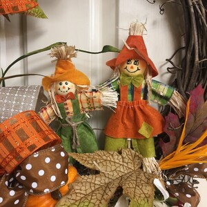 Op de afbeelding: Een herfstkrans met twee vogelverschrikkerfiguren met strohaar, oranje hoeden en groene en oranje kleding. De krans bevat pompoenen, herfstbladeren en bruin en wit stippenlint.