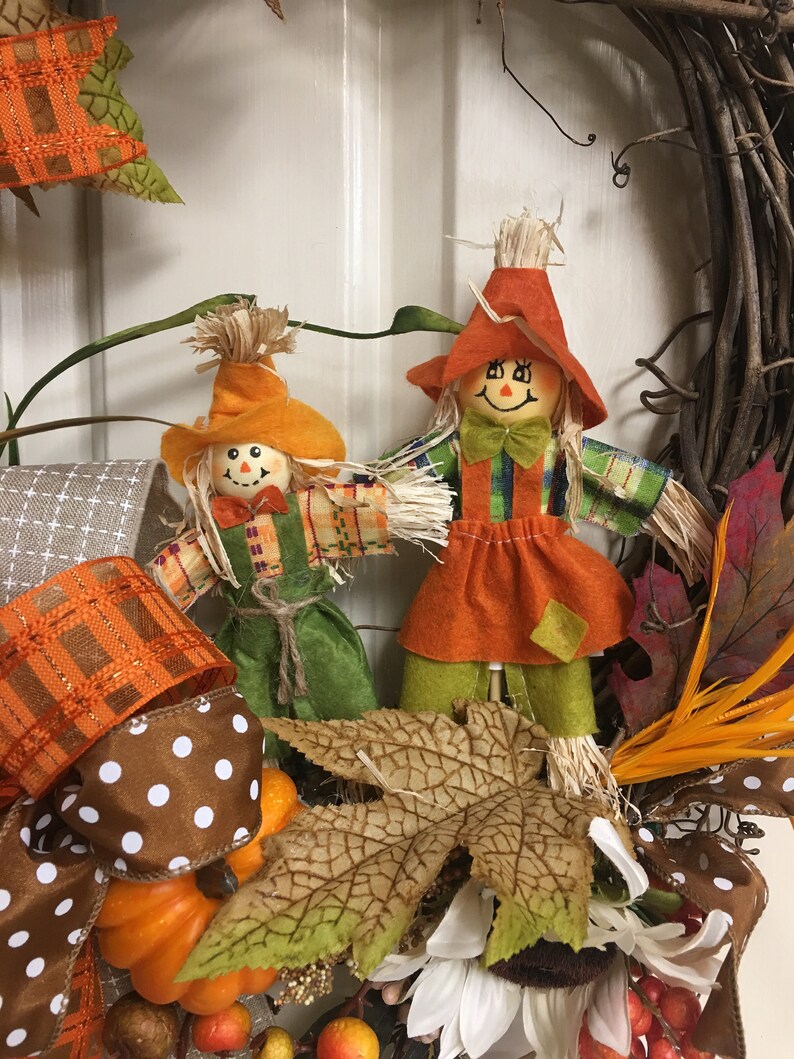 Op de afbeelding: Een herfstkrans met twee vogelverschrikkerfiguren met strohaar, oranje hoeden en groene en oranje kleding. De krans bevat pompoenen, herfstbladeren en bruin en wit stippenlint.