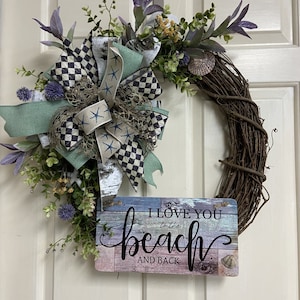 May include: A grapevine wreath decorated with a large bow featuring a black and white checkered pattern, seafoam green ribbon, and starfish accents. The wreath includes faux greenery and a sign that reads "I love you to the beach and back."