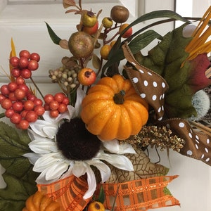 Op de afbeelding: Een herfstkrans met oranje pompoenen, witte zonnebloemen en diverse bessen. De krans bevat een bruin lint met witte stippen en een oranje geruit lint, wat een feestelijke herfstuitstalling cre&euml;ert.