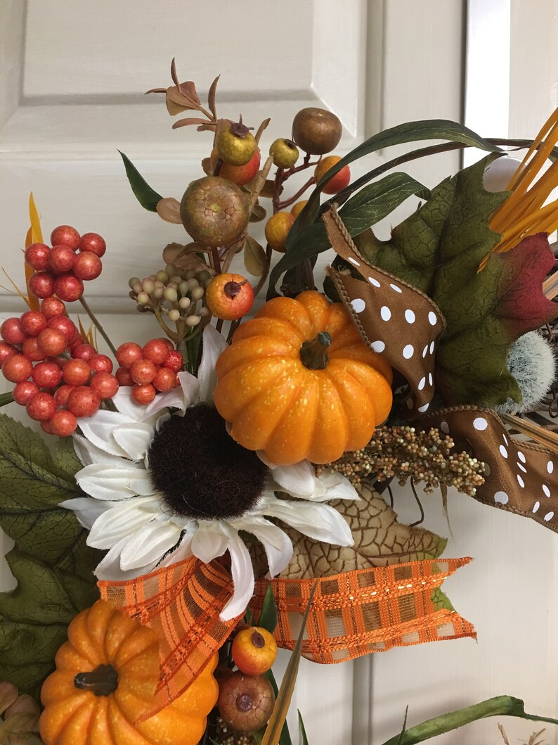 Op de afbeelding: Een herfstkrans met oranje pompoenen, witte zonnebloemen en diverse bessen. De krans bevat een bruin lint met witte stippen en een oranje geruit lint, wat een feestelijke herfstuitstalling cre&euml;ert.