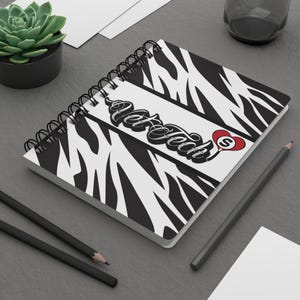 May include: A spiral-bound notebook with a zebra print design. The notebook features the text "Vet Tech" in a stylized font, with a red heart and the letter "S" on the right side. The notebook is on a gray surface.