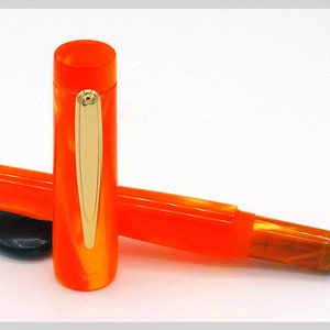 May include: A bright orange fountain pen with gold trim and a fine nib. The pen is lying on a white surface.