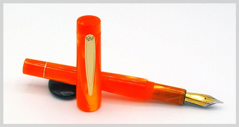 May include: A bright orange fountain pen with gold trim and a fine nib. The pen is lying on a white surface.