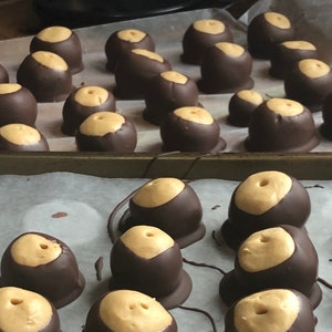 May include: A baking sheet lined with parchment paper holds rows of chocolate-covered peanut butter candies. The candies are arranged in a grid pattern and have a smooth, glossy finish.