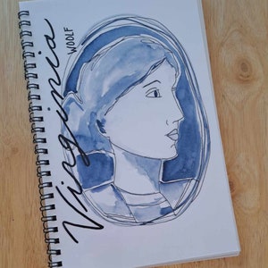 May include: A blue watercolour illustration of a person's face in a circle with the words "Virginia Woolf" written in black cursive.