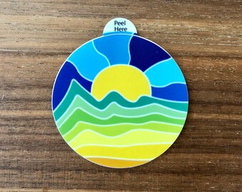 May include: A round sticker with a colorful design of a sunset over a mountain range. The sticker has a blue, green, and yellow color scheme. The text "Peel Here" is printed on the sticker.