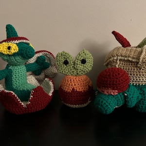 Puede incluir: Colección de figuras de ganchillo hechas a mano en varios colores. Las figuras incluyen una criatura verde y roja que emerge de un caparazón, una pequeña figura verde y naranja y una figura en forma de manzana beige, verde y roja.